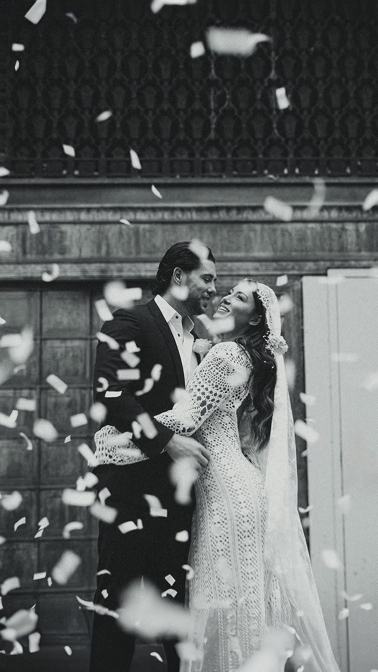 Bride and groom embracing and smiling while white confetti falls around them.