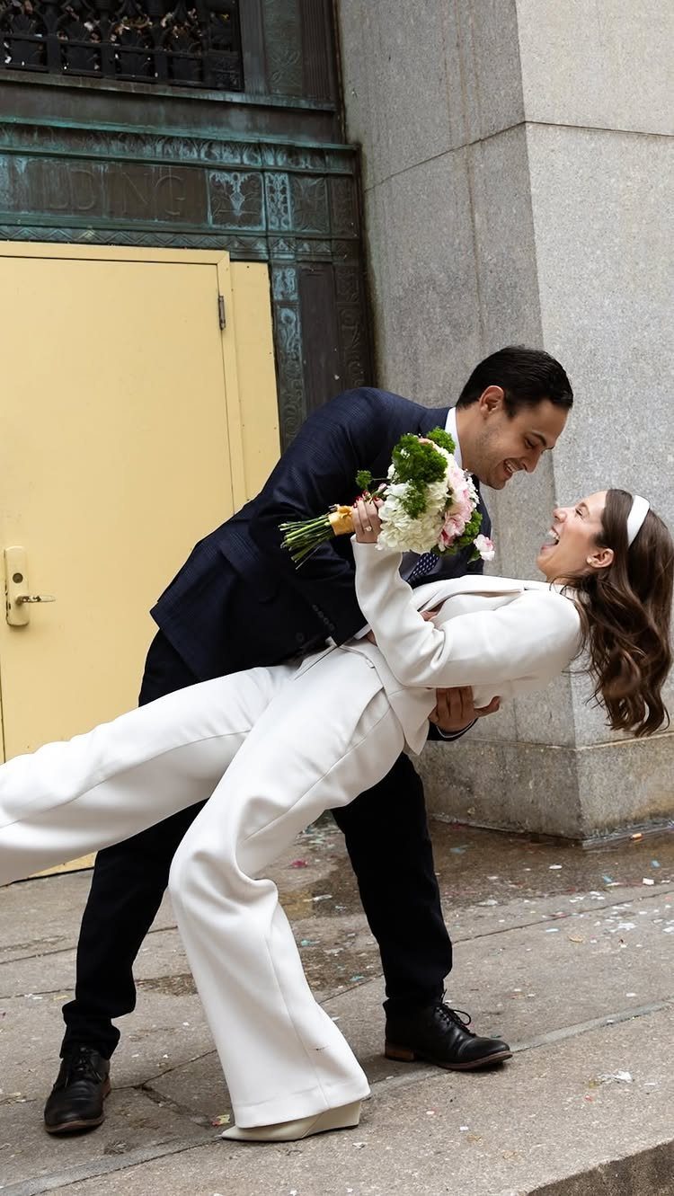 Groom in navy suit dipping bride in white pantsuit outside City Hall