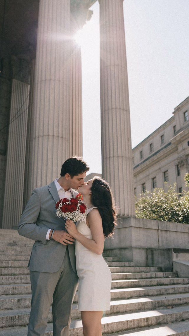 Couple kissing in front of neoclassical stone columns with a red flower bouquet