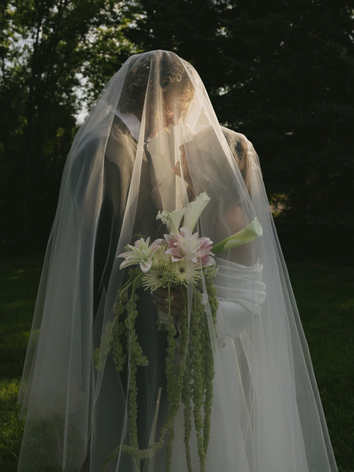 Bride and groom embracing under a sheer white veil holding a lily bouquet.