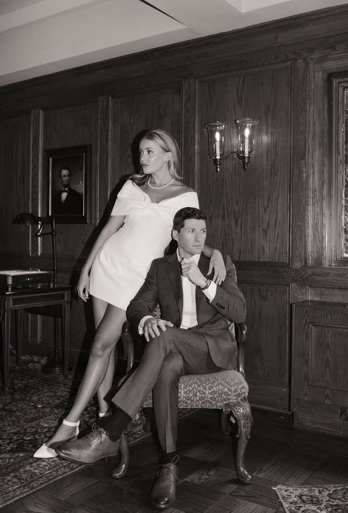 Couple posing in a wood-paneled room, woman in white dress and man in dark suit.