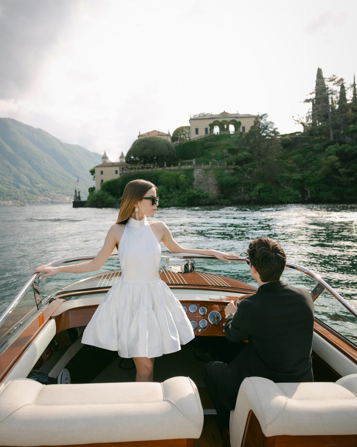 Couple on a wooden luxury boat with a grand villa and mountains behind them.