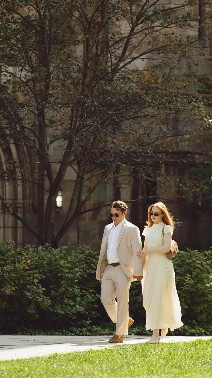 Couple in elegant beige and white attire walking hand-in-hand near a stone building.