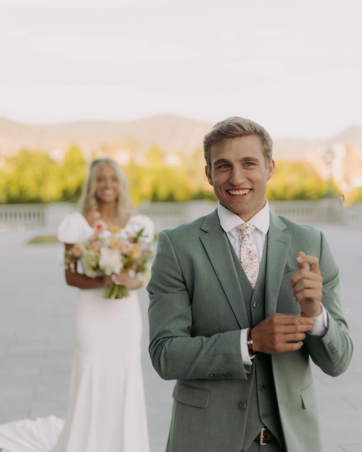 Smiling groom in green suit with bride approaching him in the blurred background