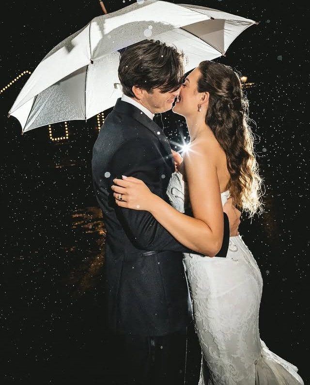 Bride and groom embracing under a white umbrella during a rainy night photoshoot.