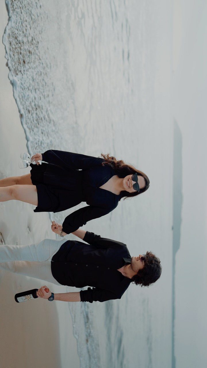 Couple in black outfits holding hands on a sandy beach by the ocean.