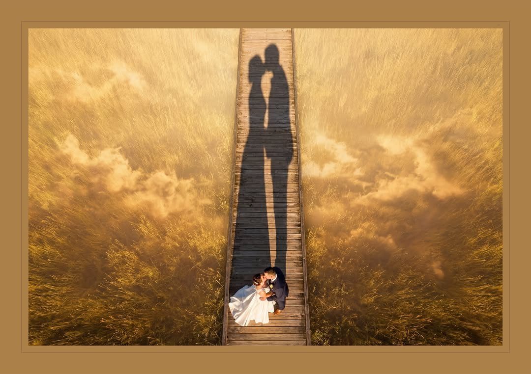 Bride and groom embracing on a wooden pier with long silhouettes stretching forward.