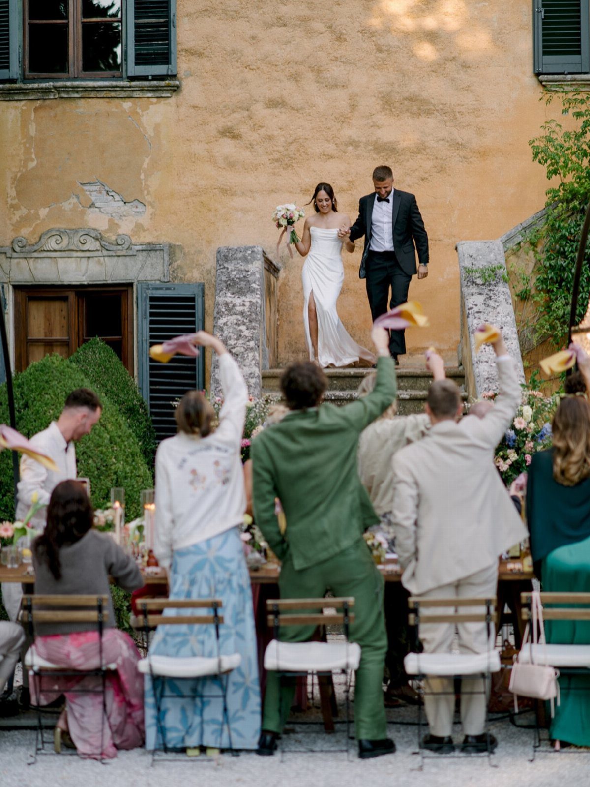 Bride and groom walking together down stone steps while guests cheer at a table.