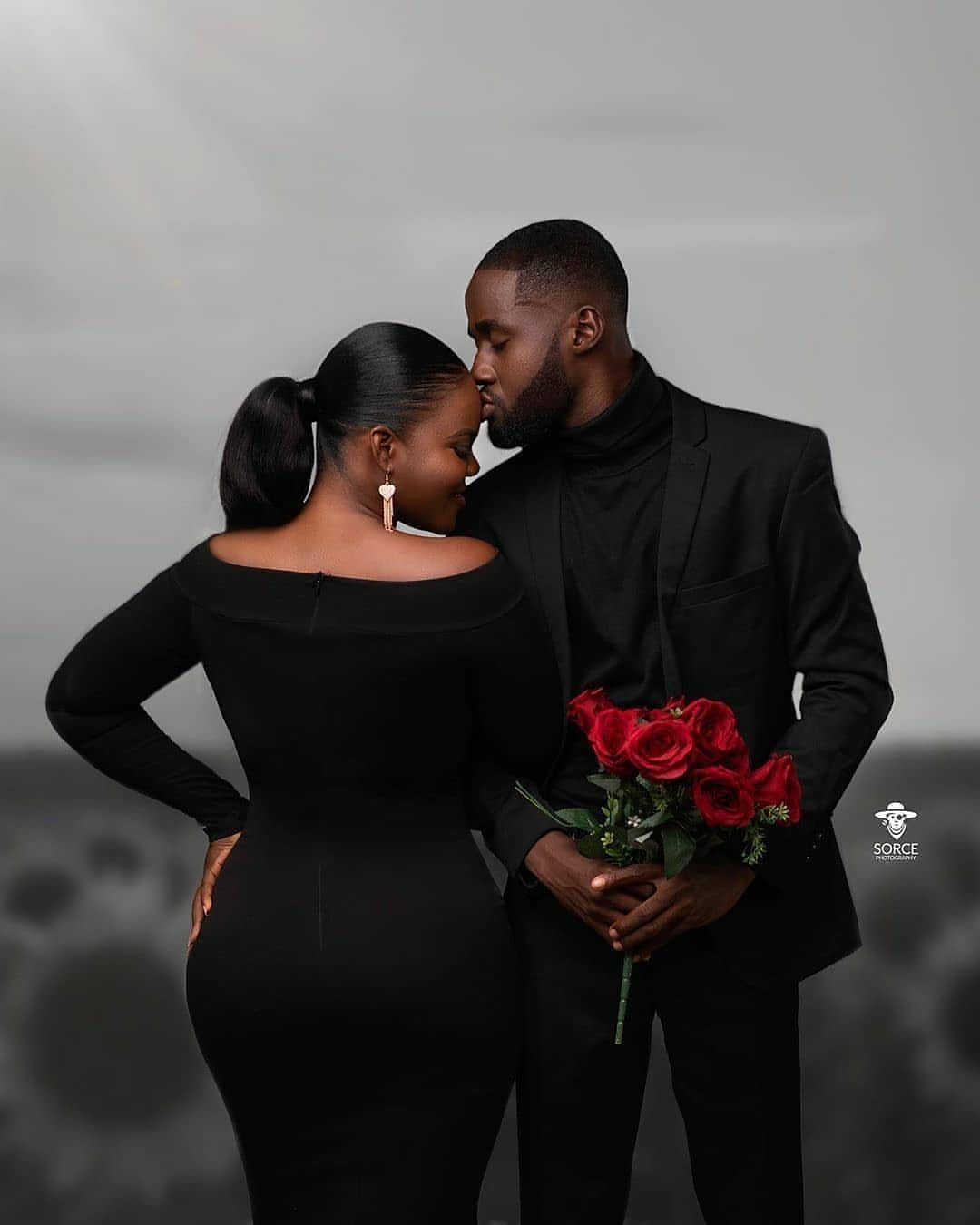 Couple in all-black outfits posing together with a bouquet of red roses.