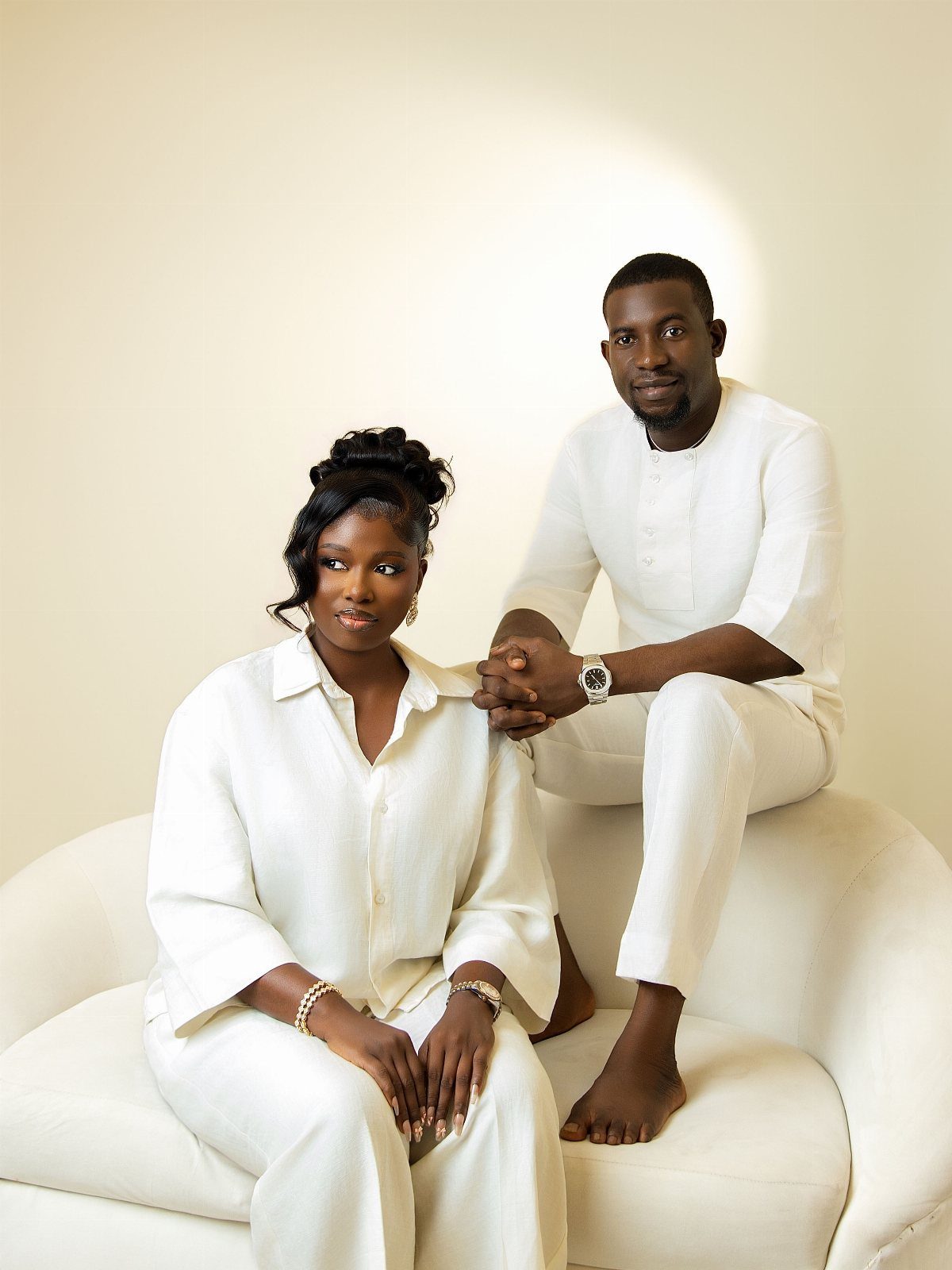 Black couple wearing matching all-white linen outfits posing on a white sofa