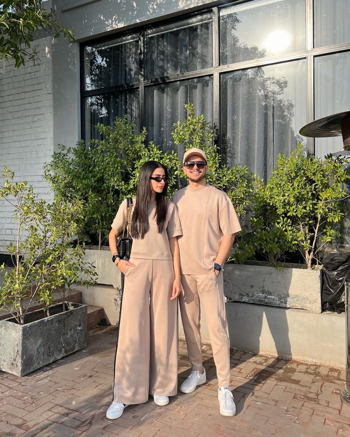 Couple wearing matching beige coordinated outfits and white sneakers standing outdoors near greenery.