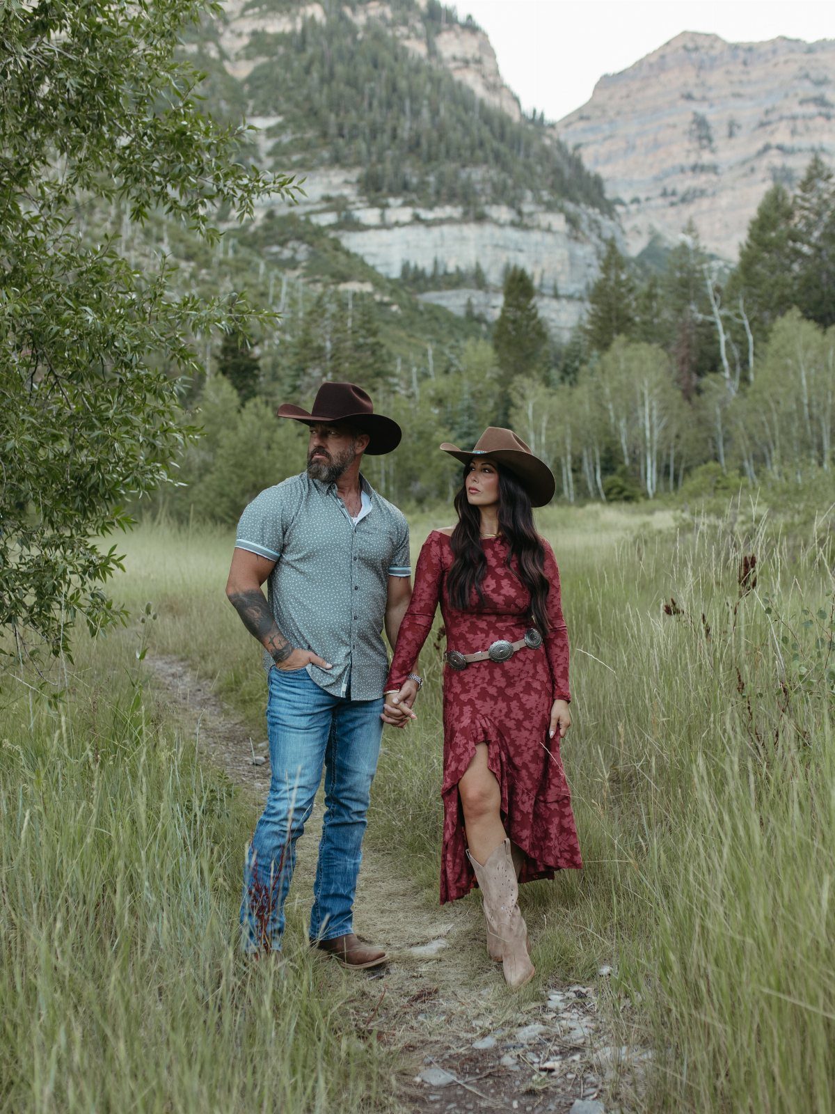 Couple in cowboy hats and western attire walking through a grassy mountain valley.