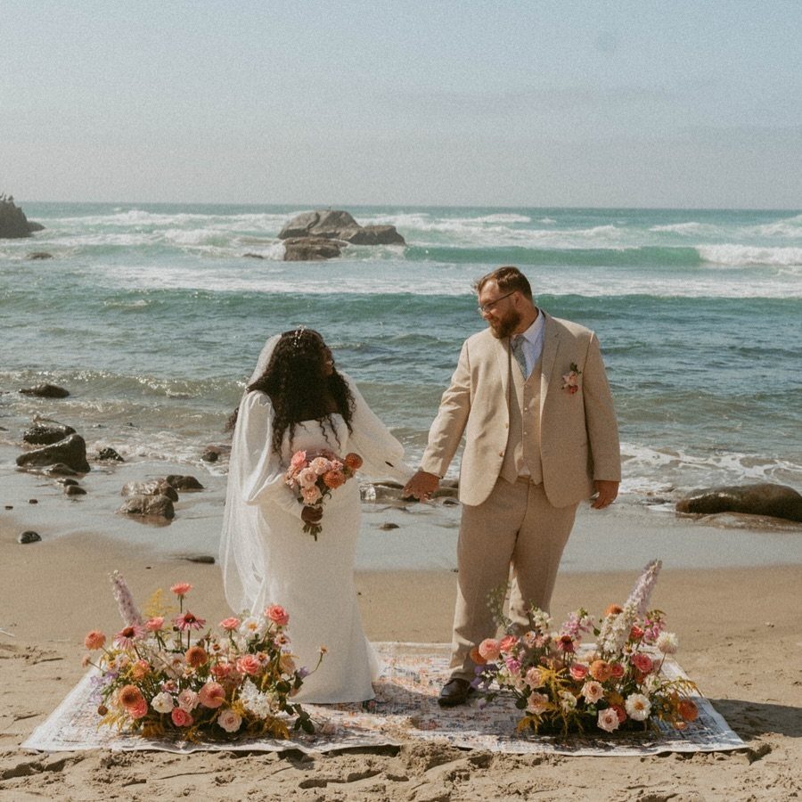 Plus-size bride and groom holding hands on a beach rug with floral arrangements.