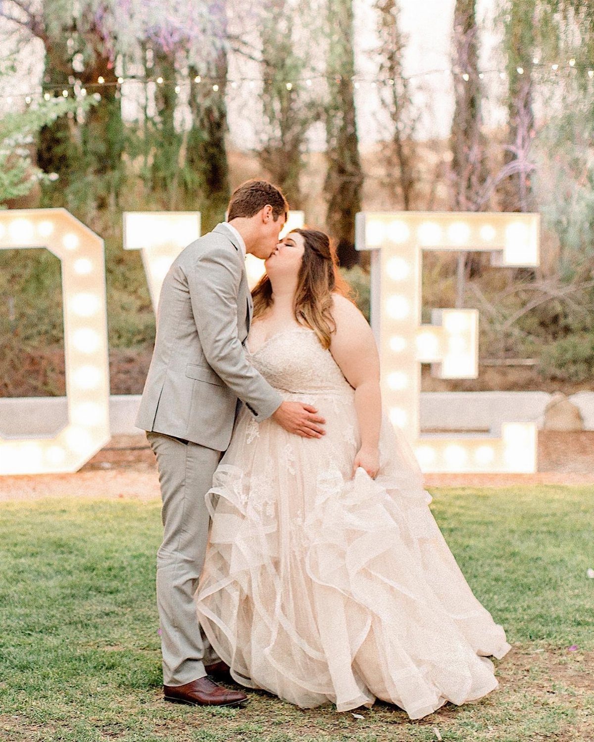 Plus-size bride in ruffled gown and groom in grey suit kissing outdoors.