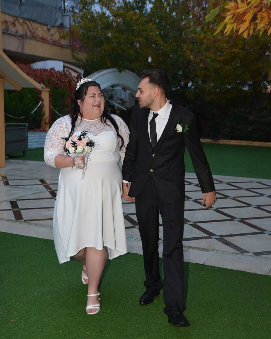 Plus-size bride in white lace dress and groom in black suit walking together.