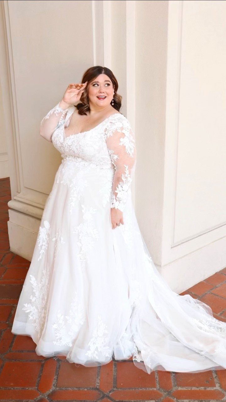 Plus-size bride in a white lace ball gown posing by a white wall.
