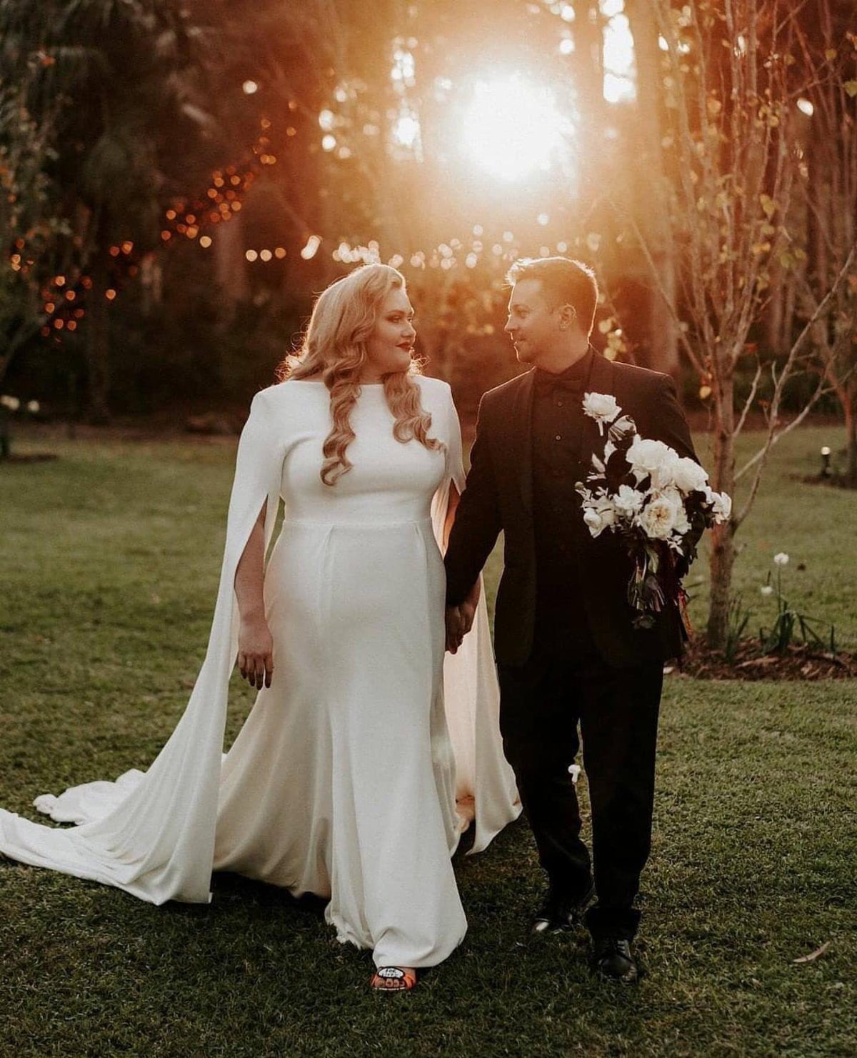 Plus-size bride and groom walking hand-in-hand in a grassy field at sunset.