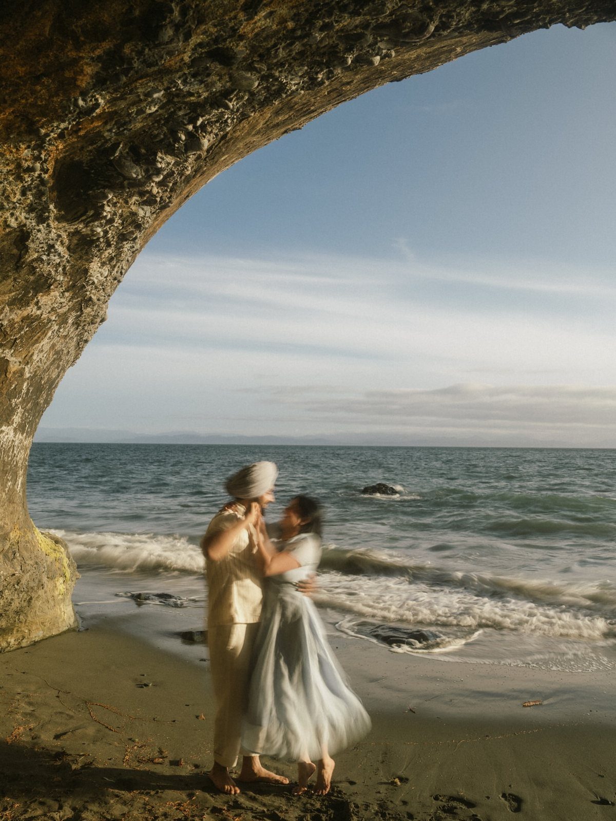 Couple embracing on a sandy beach framed by a natural rock archway.