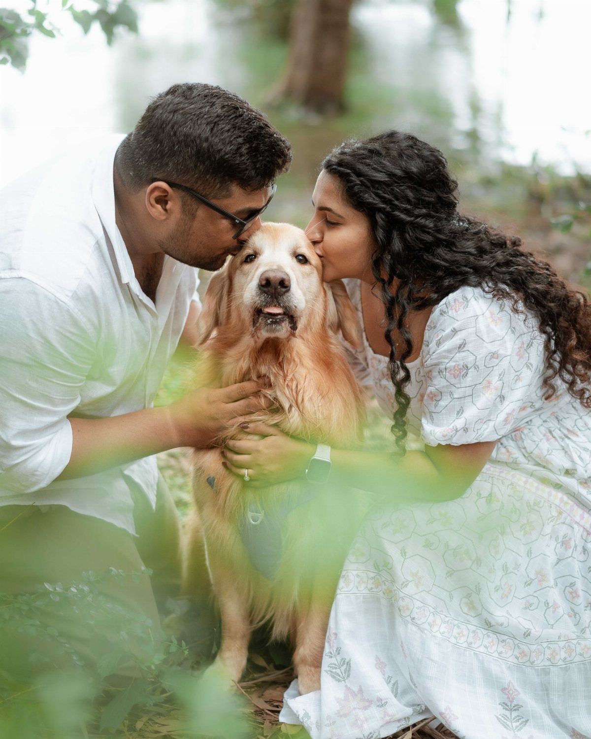 Couple in white clothing kissing their golden retriever dog in a lush green forest