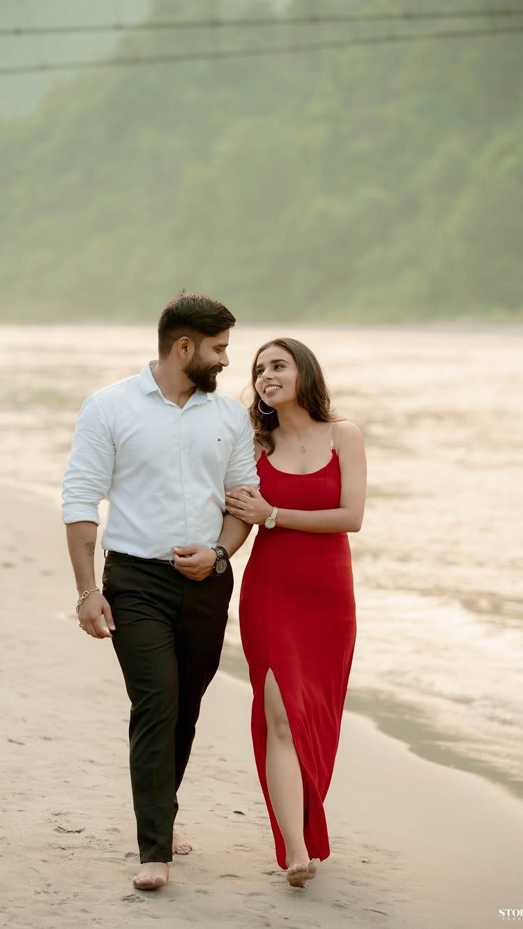Couple walking on a sandy riverbank with green hills in the background.