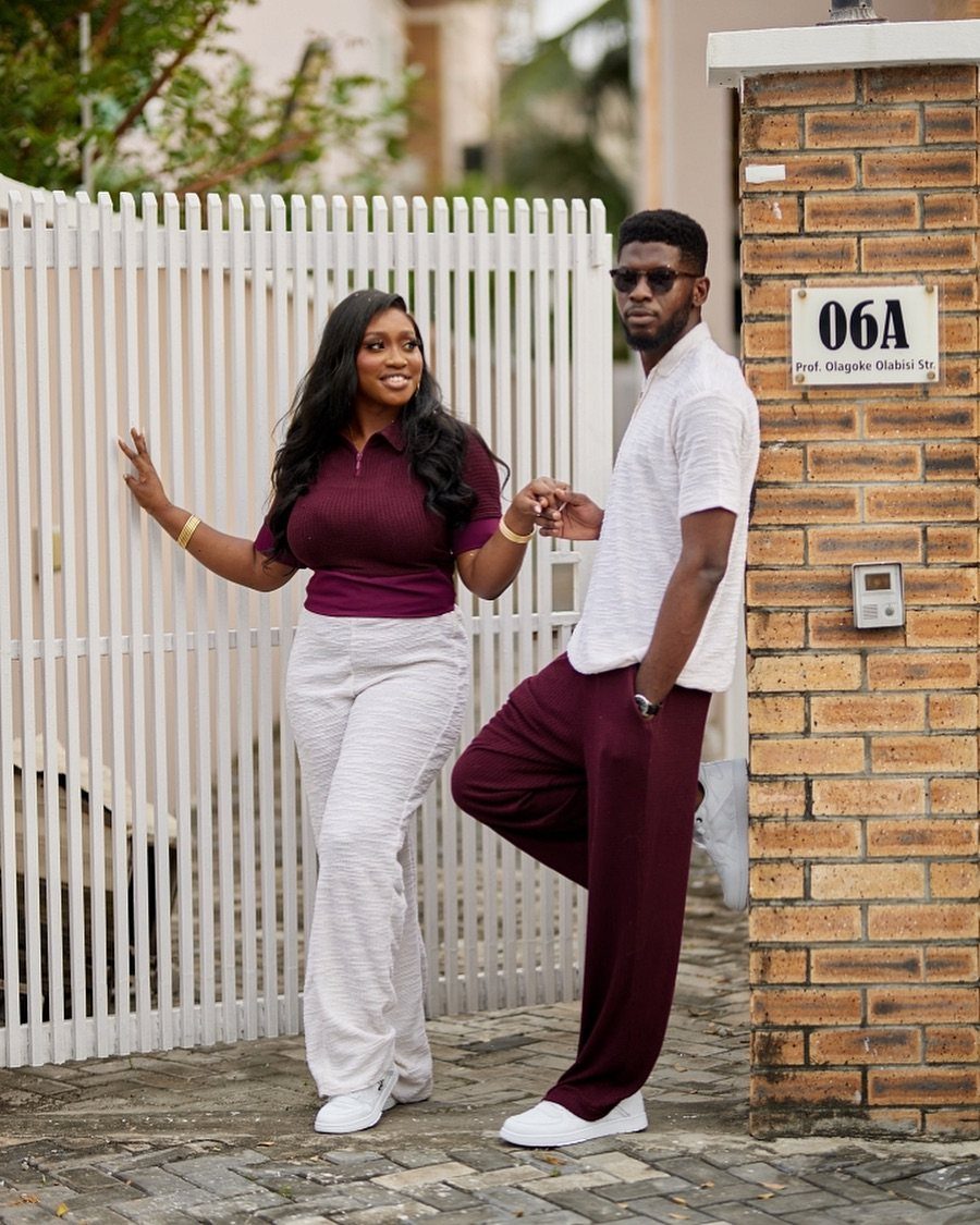Couple posing in coordinating maroon and white casual outfits by a white fence.