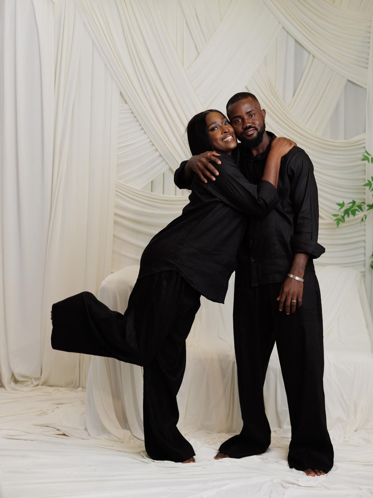 Smiling Black couple in matching oversized black outfits posing against white draped fabric.
