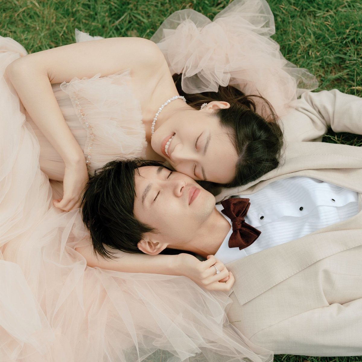 Couple lying on grass, woman in pale pink gown and man in beige suit.