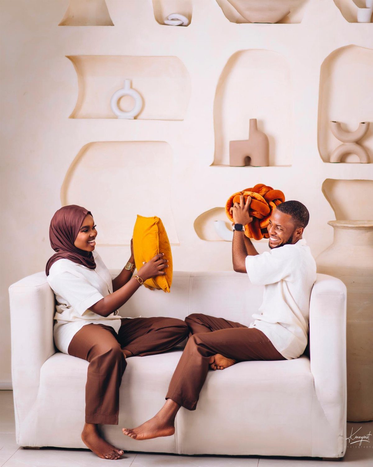 Couple in matching white shirts and brown pants laughing on a white sofa