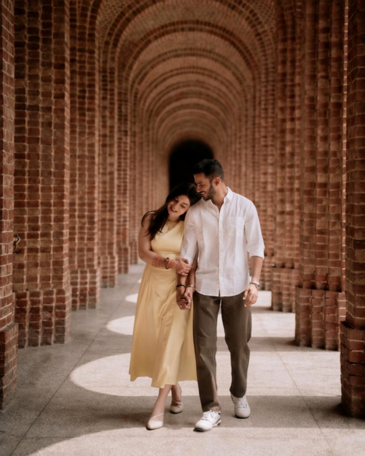Couple walking hand-in-hand through a red brick arched corridor, smiling at each other.