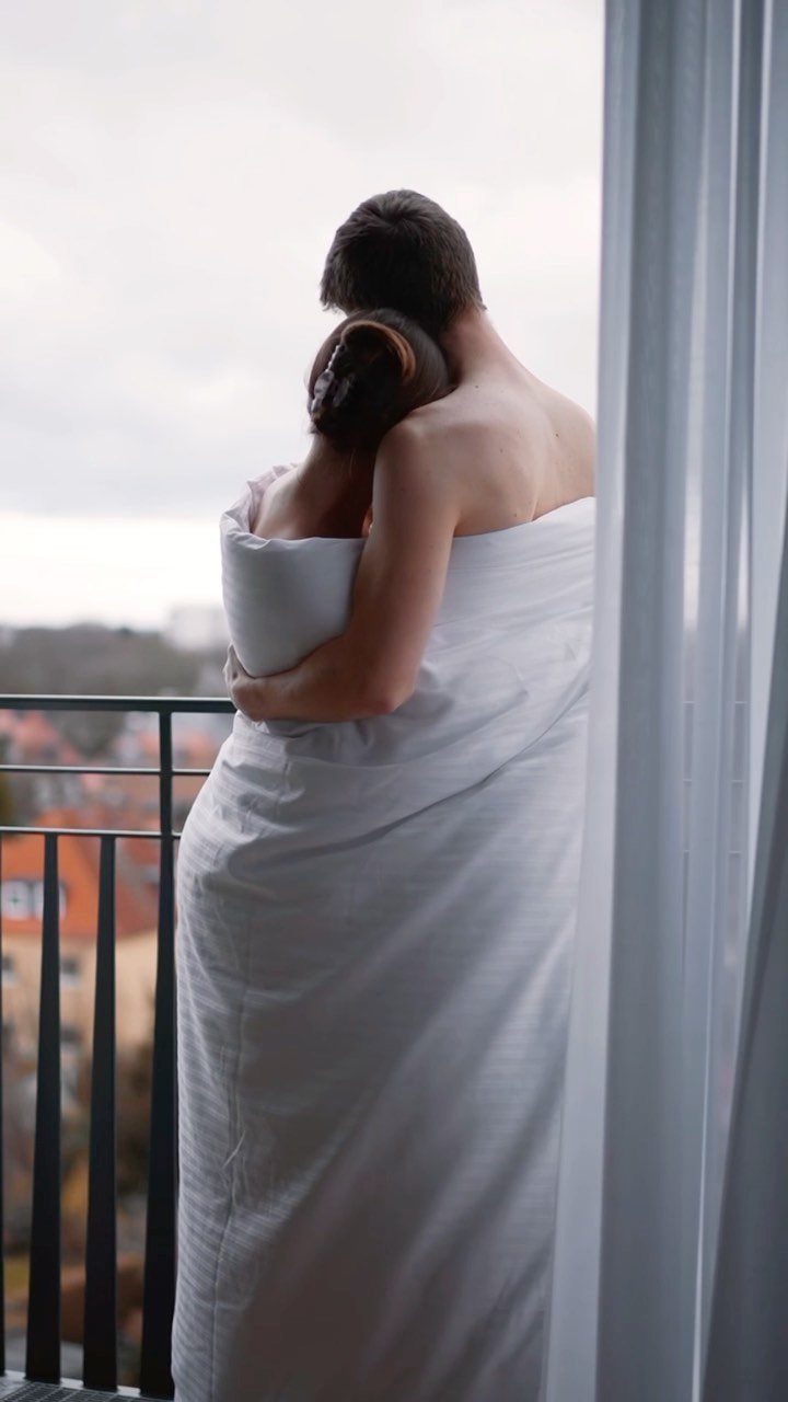 Couple wrapped in a white blanket hugging on a balcony overlooking a town.