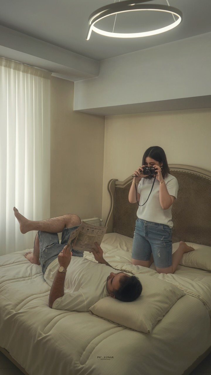 Woman photographing a man lying on a bed in a neutral bedroom setting.