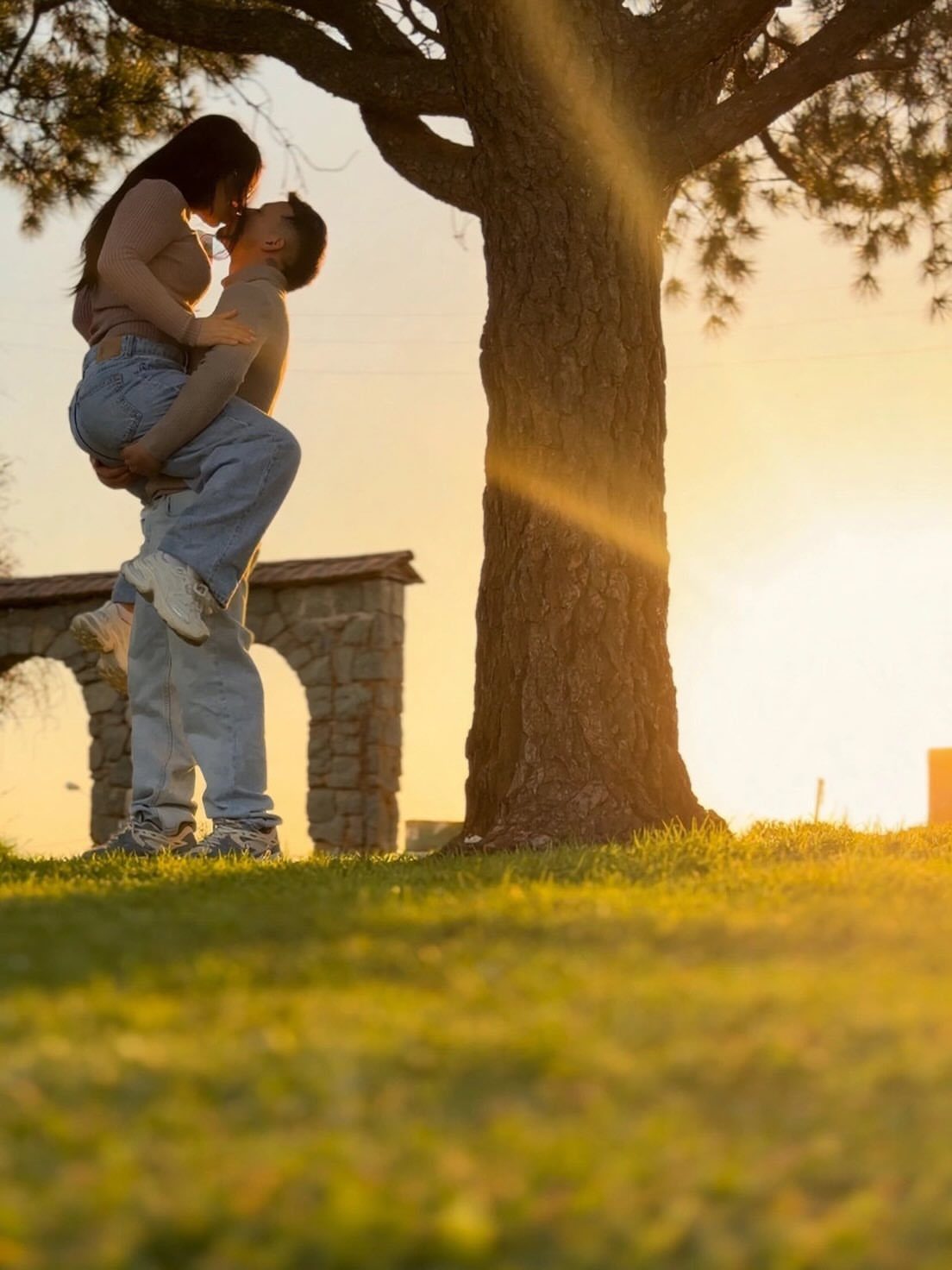 Man lifting a woman in his arms under a tree at sunset.