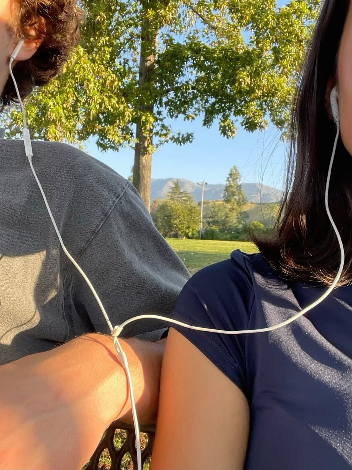 Couple sharing white wired earphones while sitting outdoors with mountains in the background