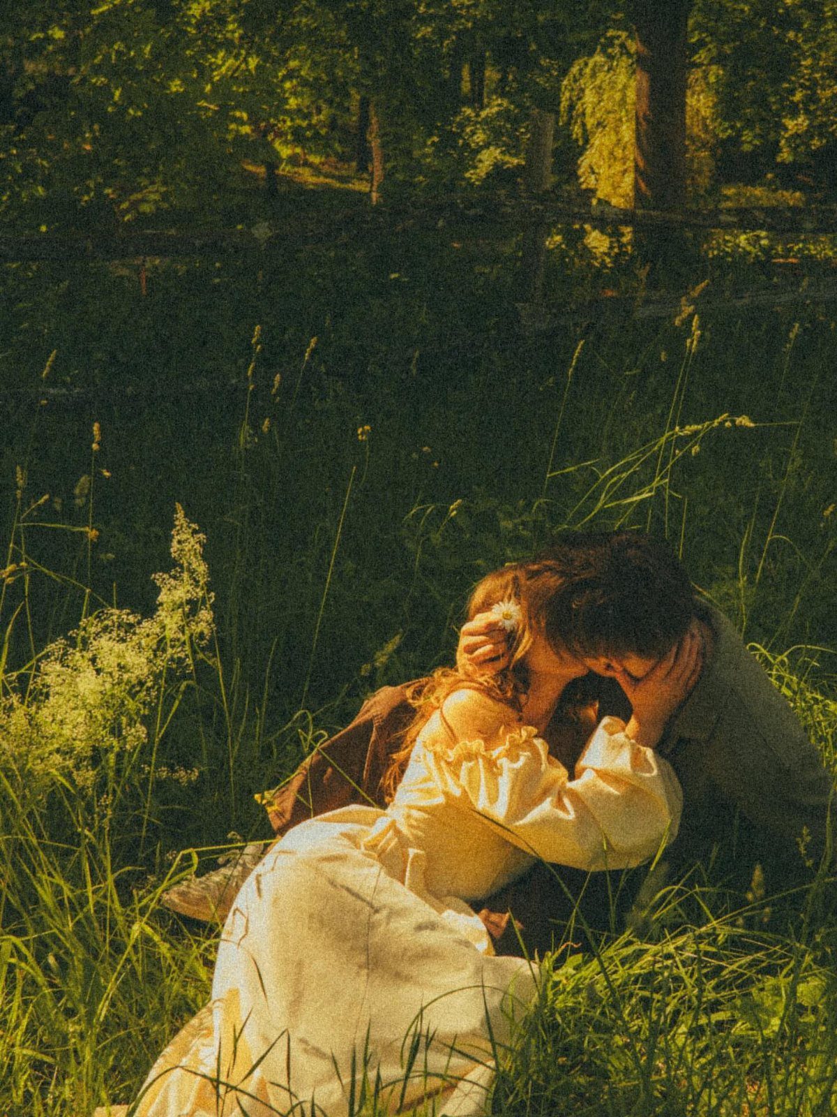 Couple in vintage attire embracing and kissing in a sunlit, grassy forest clearing.
