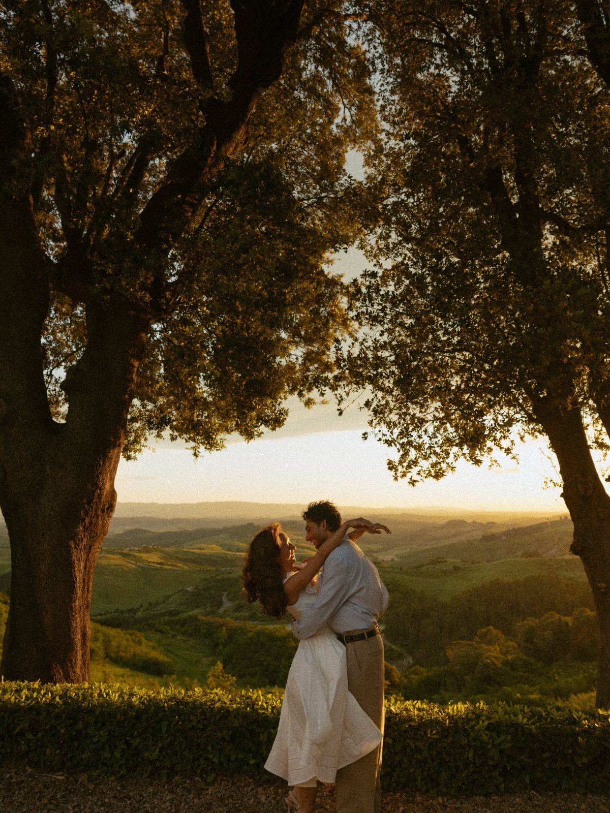 Couple embracing in a valley landscape framed by large trees at sunset.