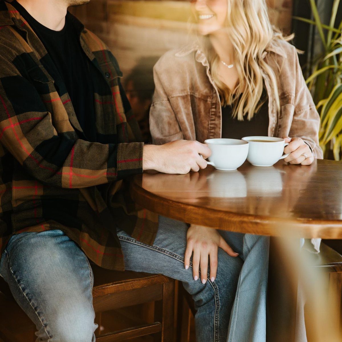 Couple sitting at a wooden table holding white coffee cups in a cafe.