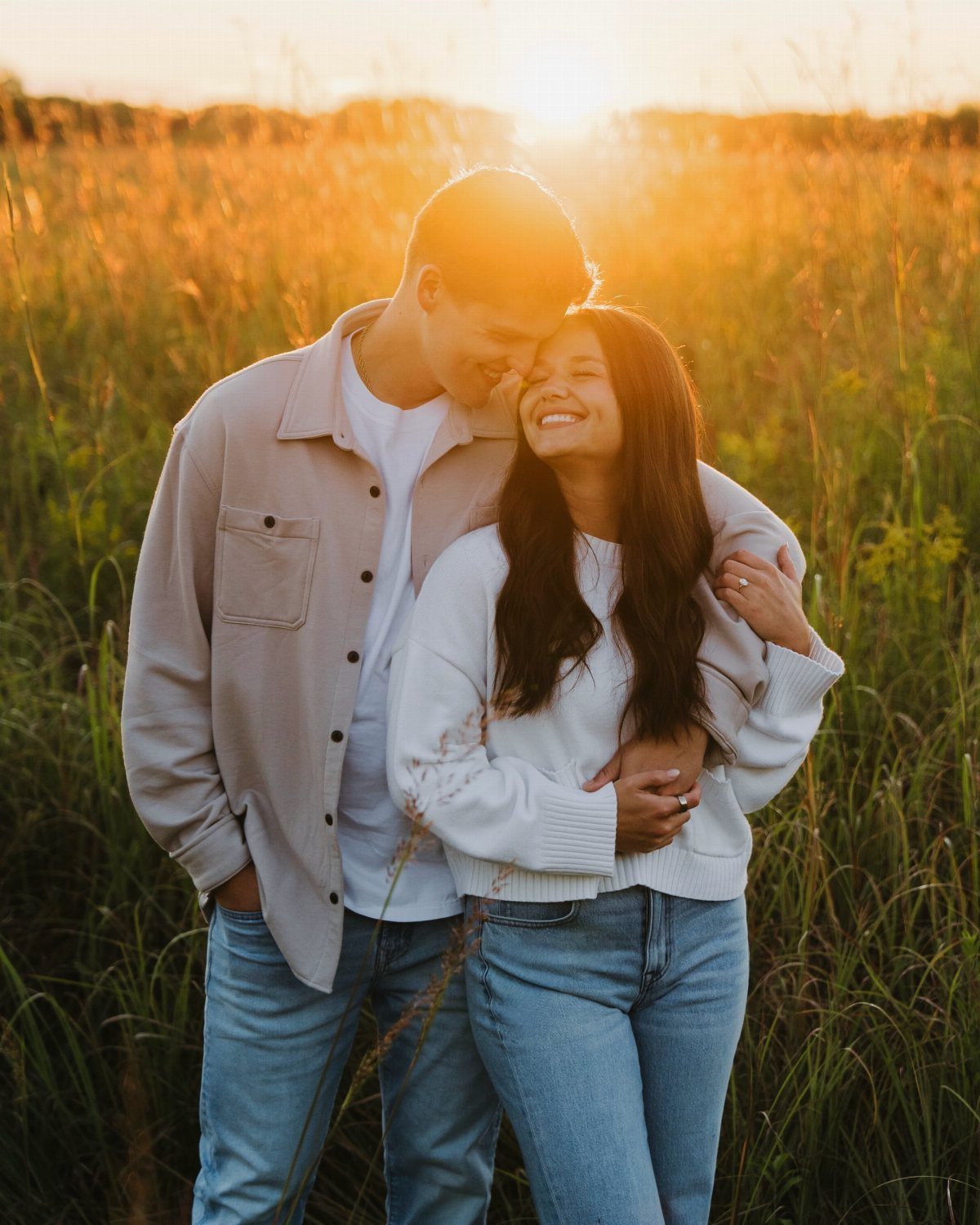 Young couple embracing and smiling in a golden grass field at sunset.