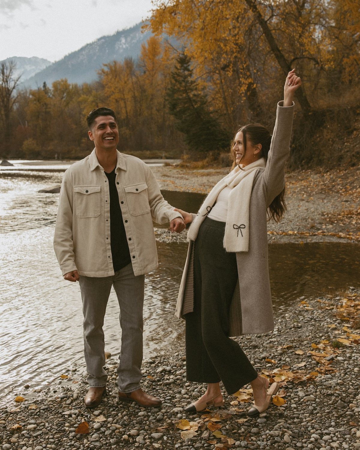 Smiling couple holding hands on a rocky riverbank surrounded by autumn foliage.