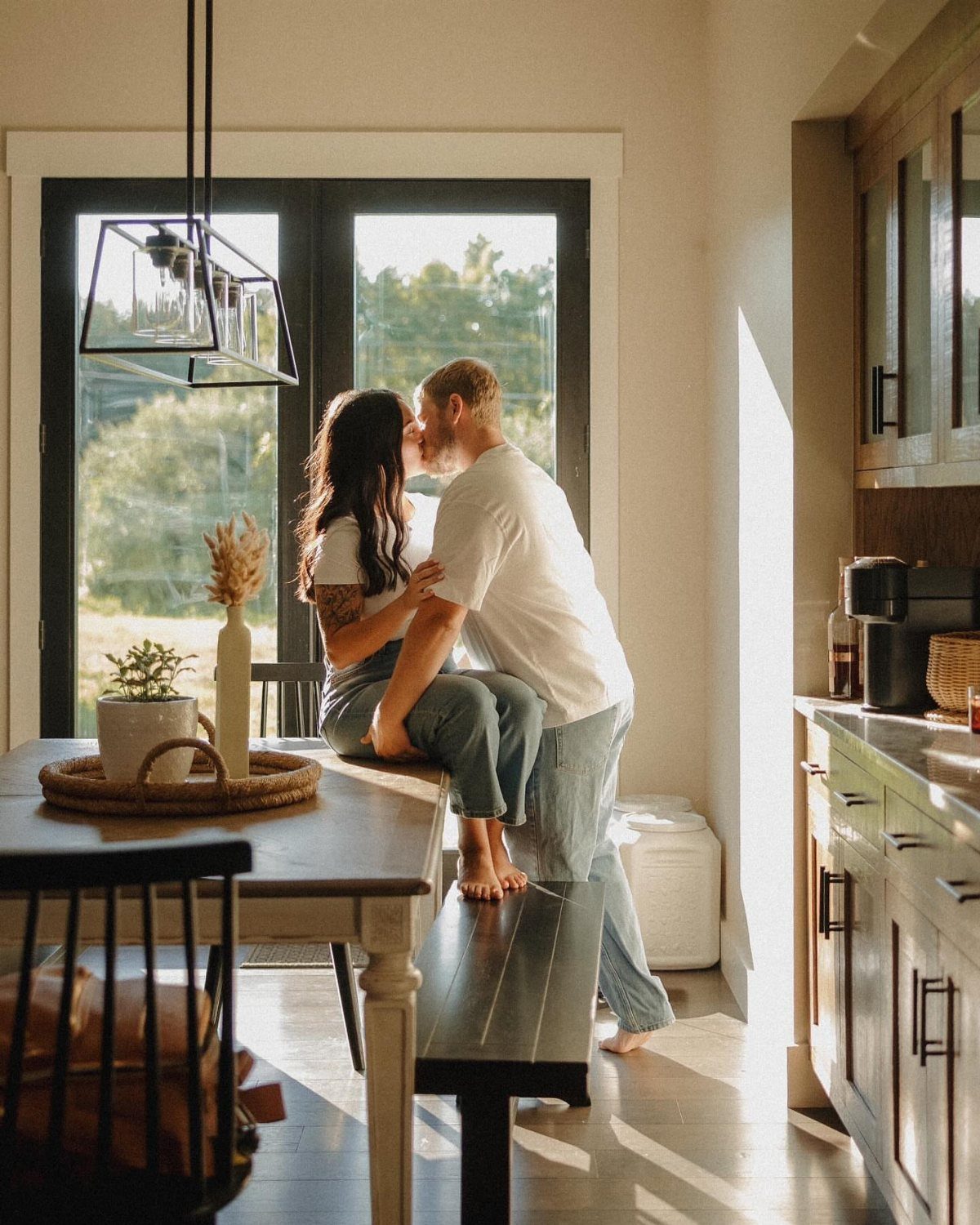 Couple kissing in a sunlit kitchen with the woman sitting on a table.