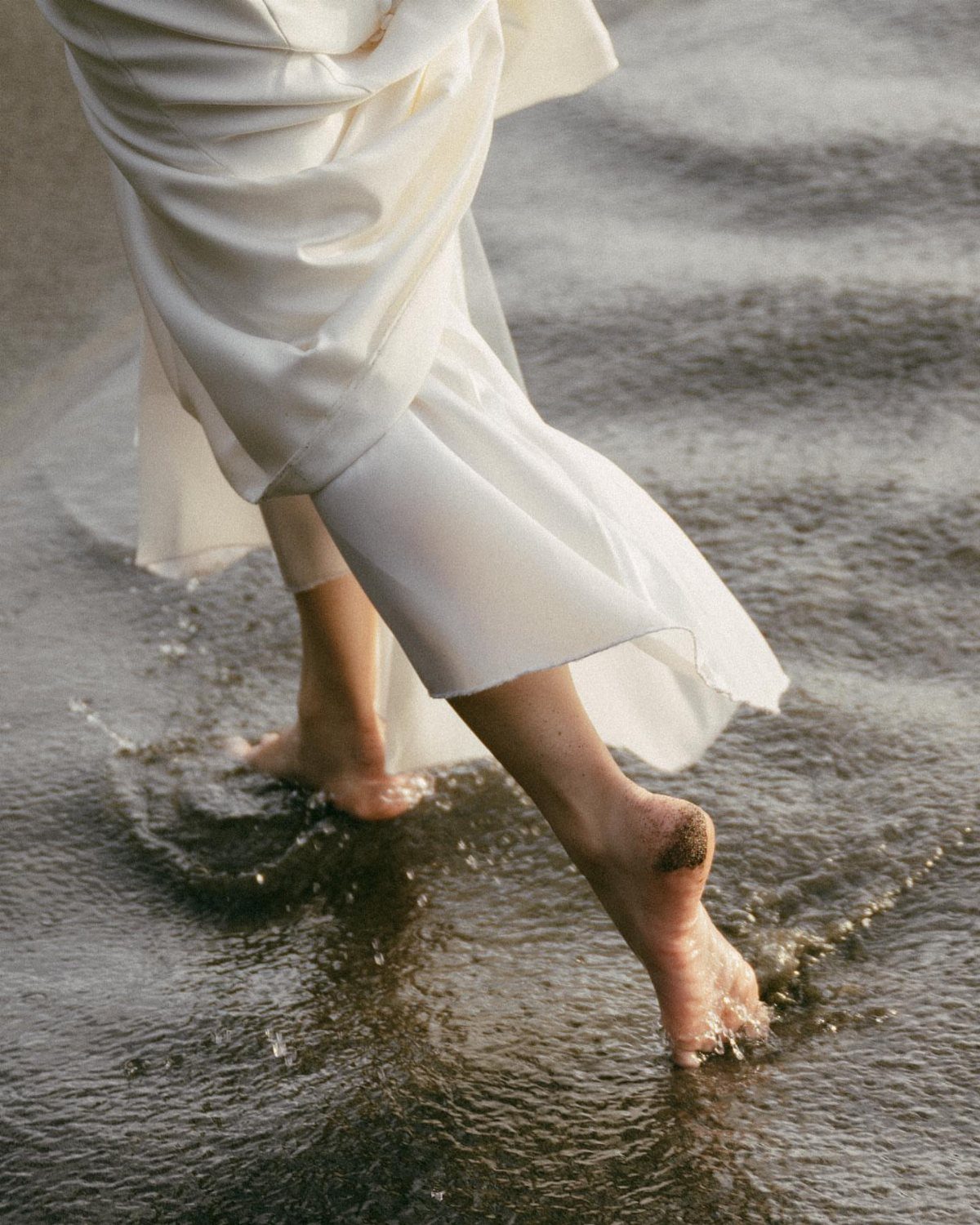 Bare feet and white dress hem walking through shallow, rippling dark water