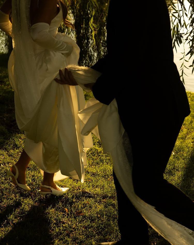 Bride in white gown and groom in black suit walking through sunny grass