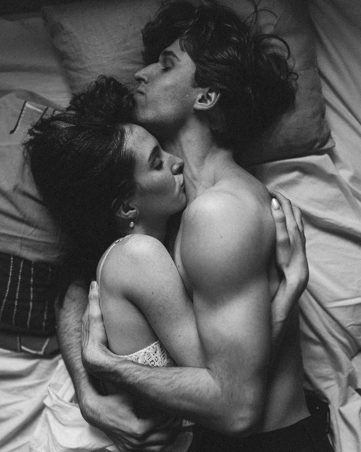 Shirtless man and woman in lace lingerie embracing intimately on a white bed.