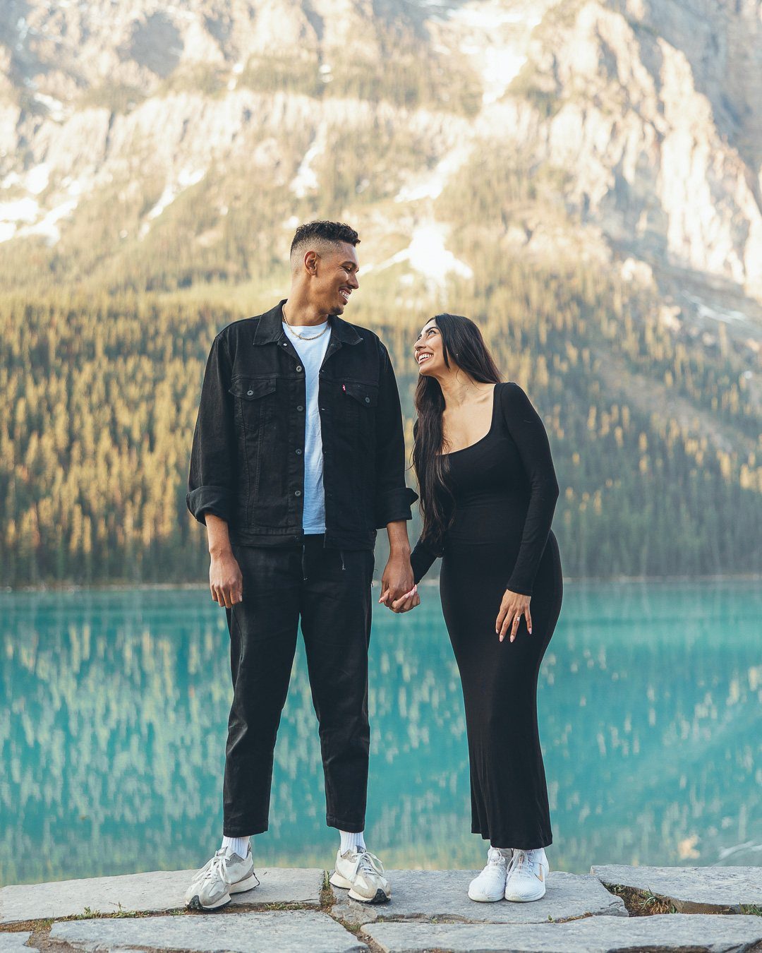 Couple in black outfits holding hands before a turquoise lake and mountains.