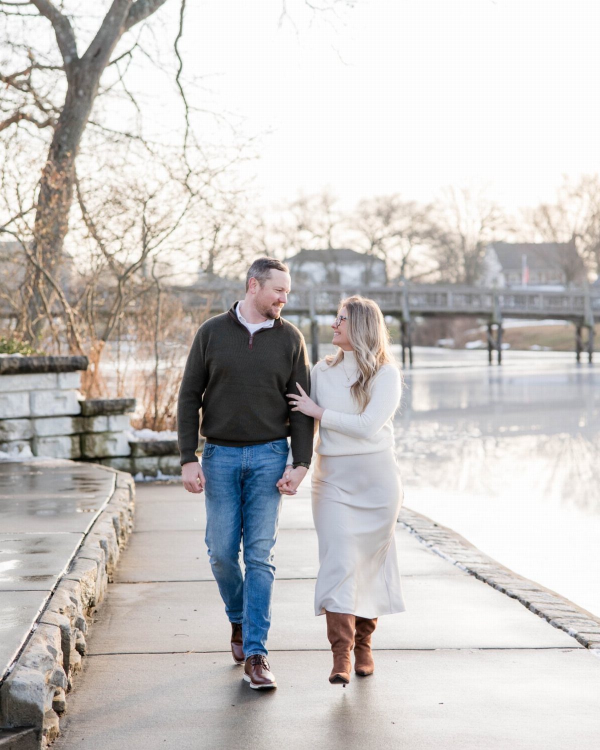 Couple walking hand-in-hand by a lake wearing neutral knit sweaters and white skirt.