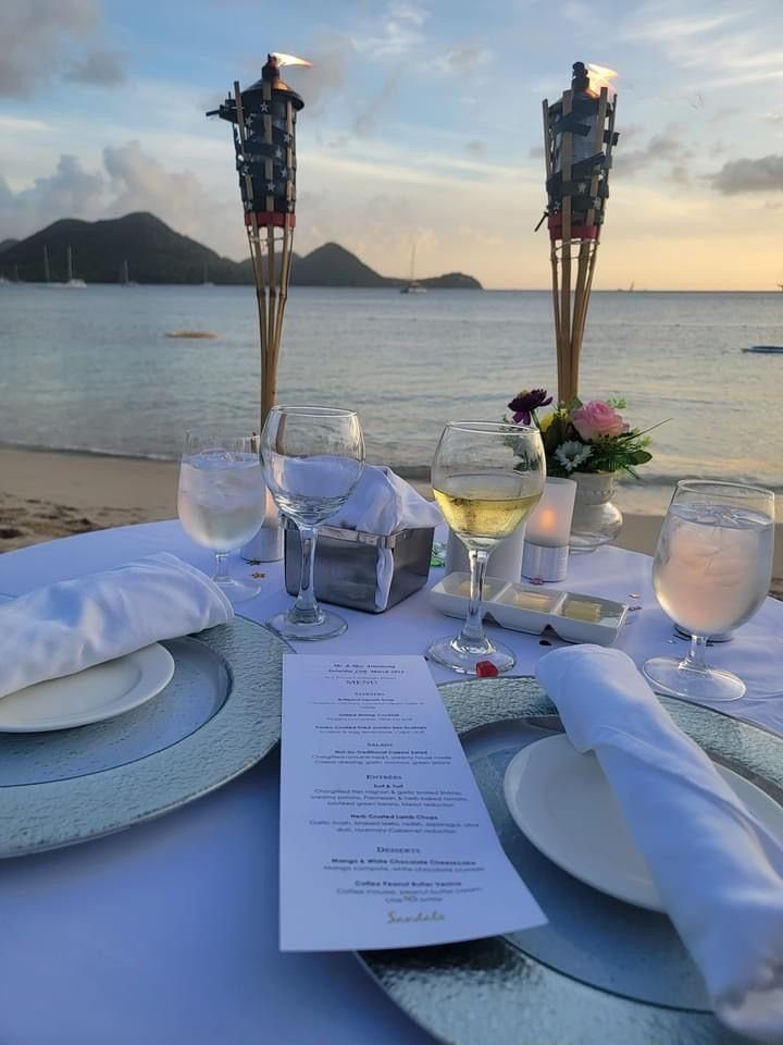 Romantic beach dinner table setting with wine glasses and tiki torches at sunset.