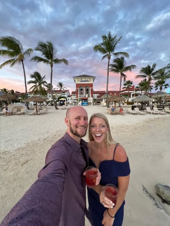 Smiling couple holding red cocktails on a tropical beach in front of a resort.
