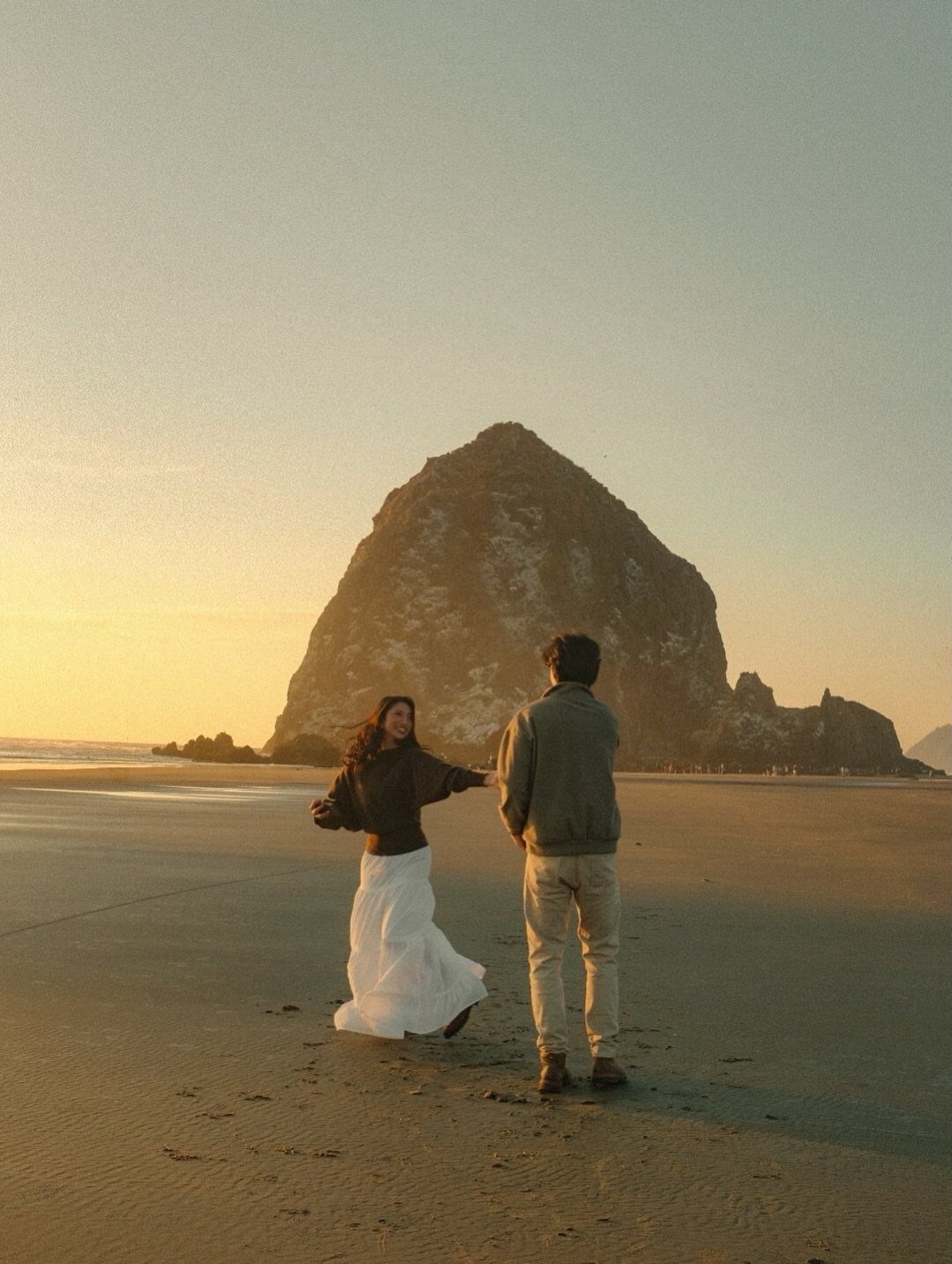 Couple holding hands on a beach with Haystack Rock in the background at sunset.