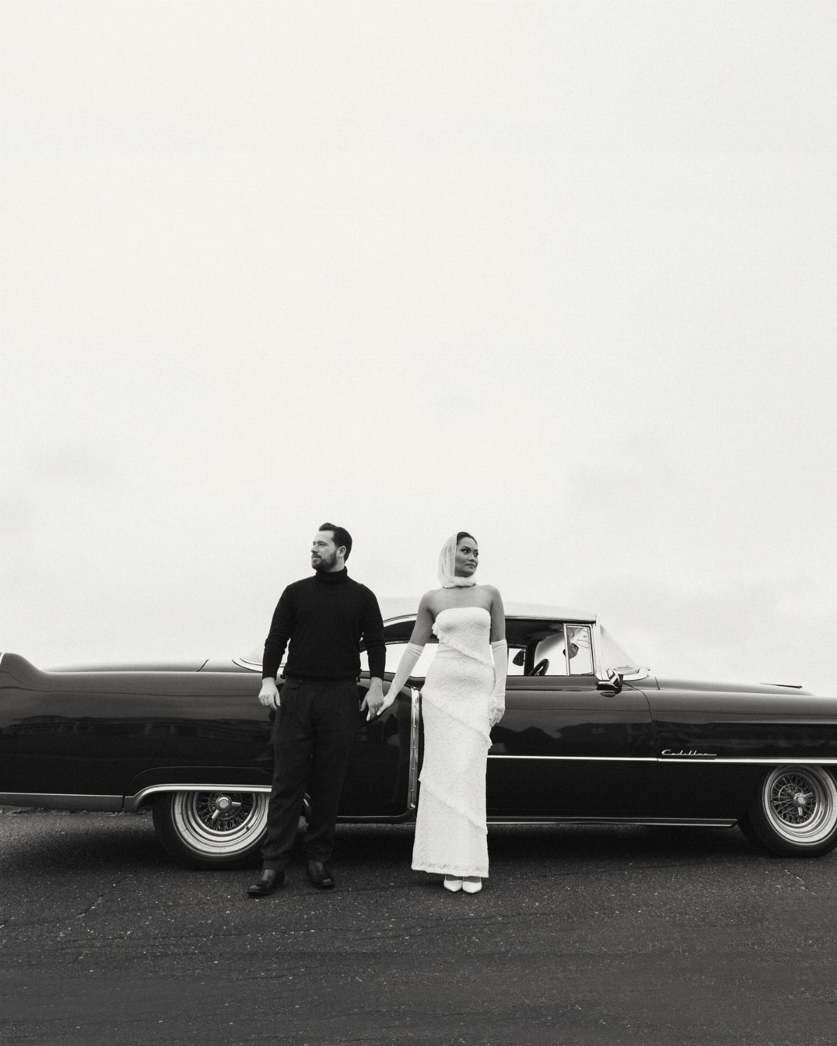 Couple in formal attire posing beside a dark vintage Cadillac in black and white.