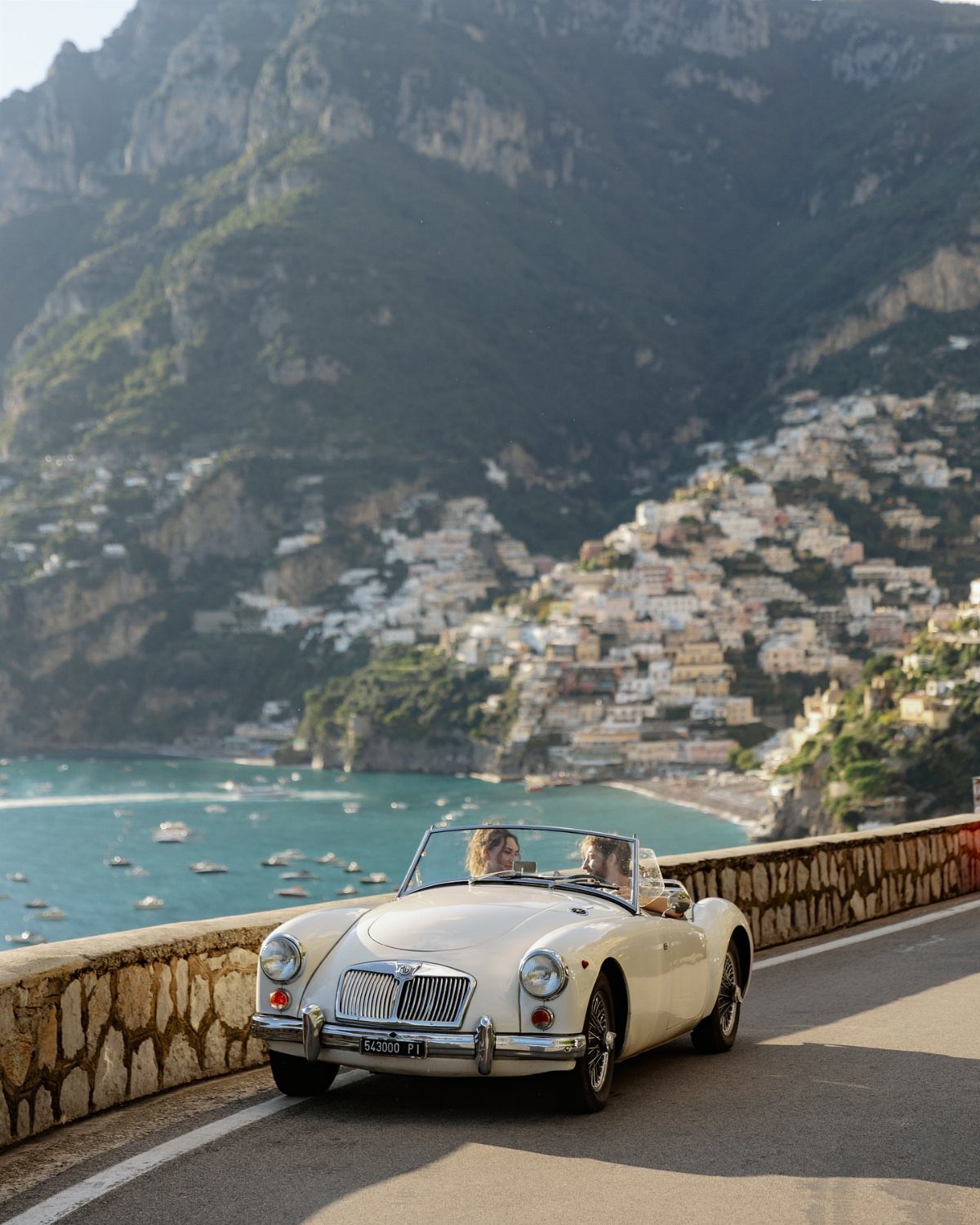 White vintage convertible driving on a coastal road overlooking a Mediterranean hillside town.