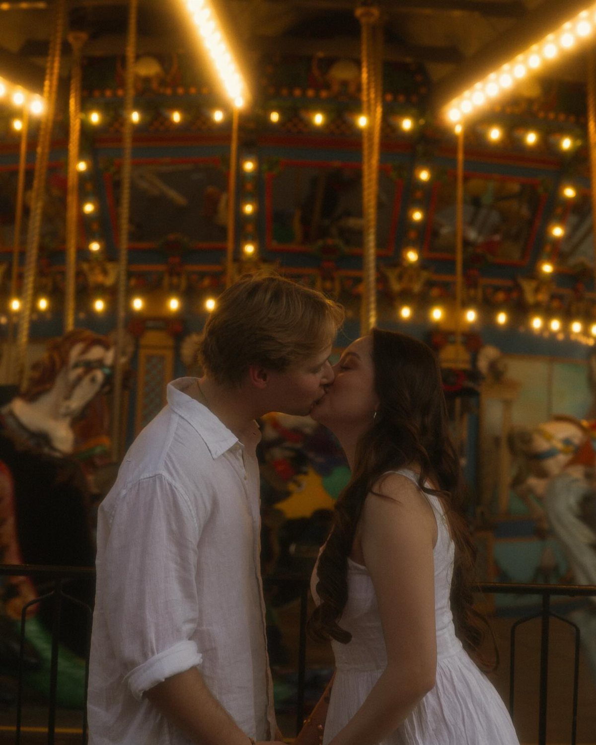 Couple kissing in front of a glowing carousel with warm yellow lights.